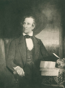 A white man in a suit sits at a desk strewn with papers. His hand is on a book.